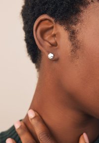Silver stud earring with a rounded, irregular shape; worn on short, curly hair near the ear; smooth surface and polished finish.