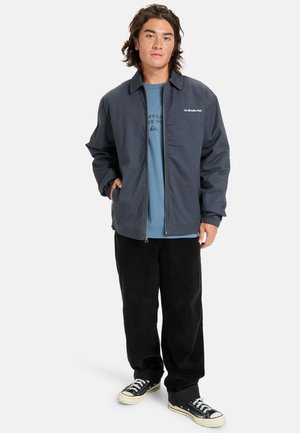 Marineblauwe jas met een ritssluiting aan de voorkant, drukknoopkraag en elastische manchetten, gedragen over een lichtblauw shirt en gecombineerd met zwarte corduroy broek en sneakers.