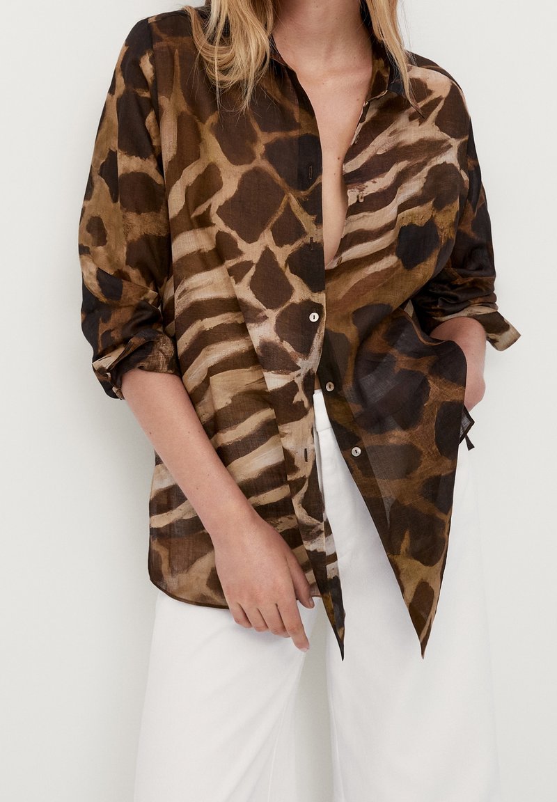Chemise ample marron et noire à imprimé animalier, boutonnée, manches retroussées, portée avec un pantalon blanc, main posée négligemment dans la poche.