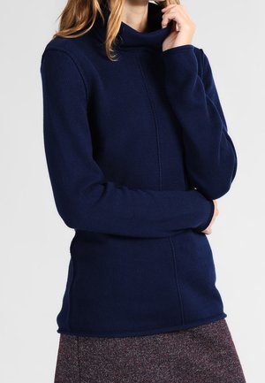 Kobieta ubrana w granatowy sweter z golfem i długim rękawem oraz brązową, teksturowaną spódnicę, jedną ręką dotykająca swojego szyi.