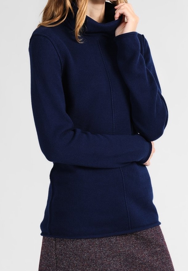 Femme portant un pull à col roulé bleu marine à manches longues et une jupe marron texturée, une main touchant son cou.