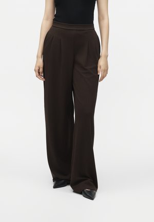 Vrouw met een hoge taille, wijde donkerbruine plooibroek, gecombineerd met zwarte puntige schoenen en een mouwloze zwarte top.