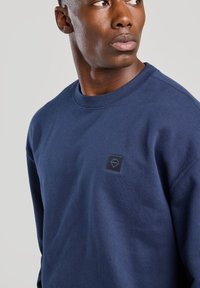 Navy blue sweatshirt in cotton material, featuring a round neckline and a square logo patch on the left chest. Smooth texture.