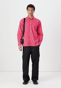Röd och vitrandig skjorta med knappar, långa ärmar, bärs med svarta cargo-byxor och en svart axelväska. Svarta skor fulländar outfiten.