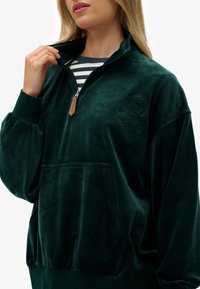Sudadera de cremallera de terciopelo verde oscuro con un alto cuello, bolsillo frontal y logotipo bordado. Presenta puños acanalados y un ajuste relajado.