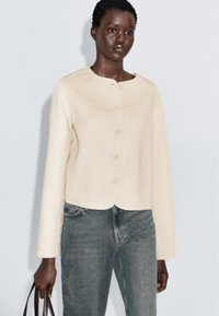 Vrouw in een crèmekleurige geknoopte cardigan en vervaagde blauwe jeans, met een bruine handtas op een effen lichte achtergrond.