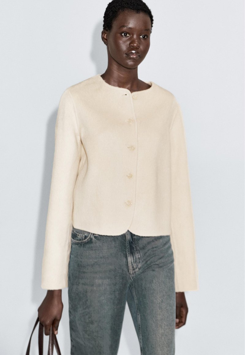 Vrouw in een crèmekleurige geknoopte cardigan en vervaagde blauwe jeans, met een bruine handtas op een effen lichte achtergrond.