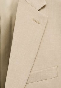 Beżowa marynarka o teksturowanej tkaninie, z lekkim kołnierzem, kieszenią na piersi oraz małym detalem w postaci przeszycia na kołnierzu.