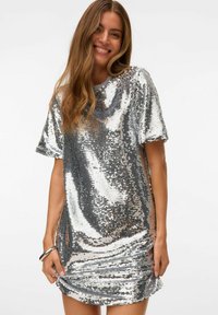 Vestito corto in paillettes argento con maniche corte, vestibilità rilassata e orlo leggermente arricciato. La texture lucida riflette la luce, aggiungendo interesse visivo.