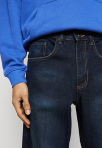 Jeans de mezclilla azul oscuro con detalles de costura clara, combinados con un sudadera azul. Los jeans presentan bolsillos estándar y cierre de botón.
