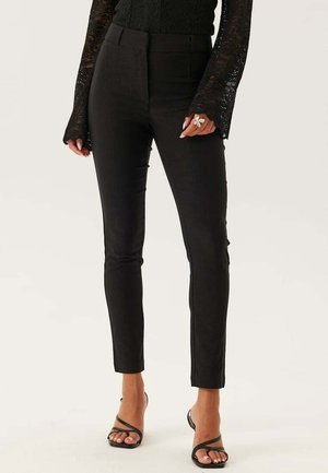 Femme portant un pantalon noir slim, un haut en dentelle noire à manches longues, des sandales à talons noires à lanières, et une grande bague papillon en argent à la main droite.