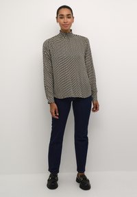 Langærmet bluse i sort og hvidt mønster med høj hals, parret med mørkeblå jeans og sorte sko. Glat stoftekstur.
