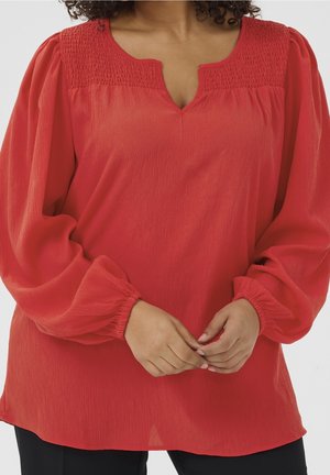 Red long-sleeve blouse with smocked yoke and gathered sleeves, worn by a person with hands clasped at the waist.