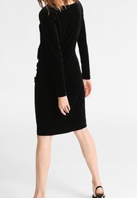 Robe en velours noir à manches longues, silhouette ajustée et ourlet à hauteur du genou. Texture lisse et design minimaliste.