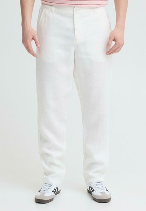 Persoon die witte, recht gesneden broek draagt, een roze gestreept overhemd in de broek gestopt, en witte sneakers met zwarte strepen, staand tegen een effen achtergrond.