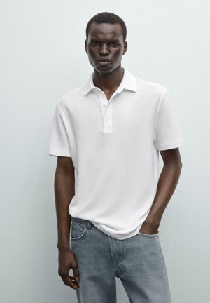Weißes Polohemd mit kurzen Ärmeln, Kragen und drei Knöpfen, kombiniert mit hellgrauen Denim-Jeans. Glatte Textur, schlichtes Design.