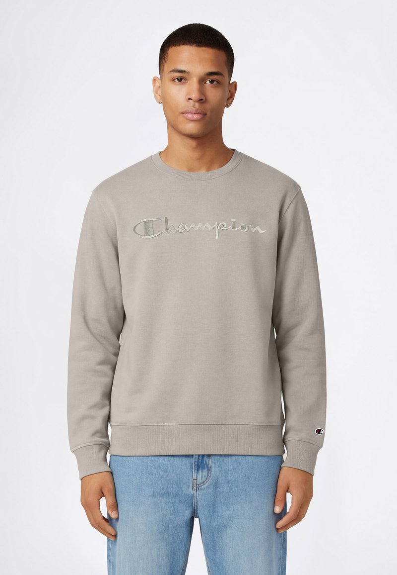 Sudadera gris con puños y dobladillo de canalé, con un logo bordado de "Champion" en el pecho. Combinada con vaqueros azul claro.