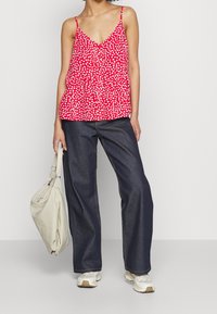 Donna che indossa un top rosso senza maniche con pois bianchi, jeans larghi scuri, sneakers bianche e tiene in mano una borsa beige.