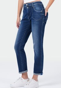 Femme portant un jean en denim bleu foncé retroussé et des baskets blanches, se tenant avec une main dans sa poche.