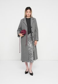 Grijze wollen jas met knopen, zwarte gebreide top, zilveren paillettenrok en een bordeauxrode trekkoordtas. Zwarte puntige ballerina's maken de look af.