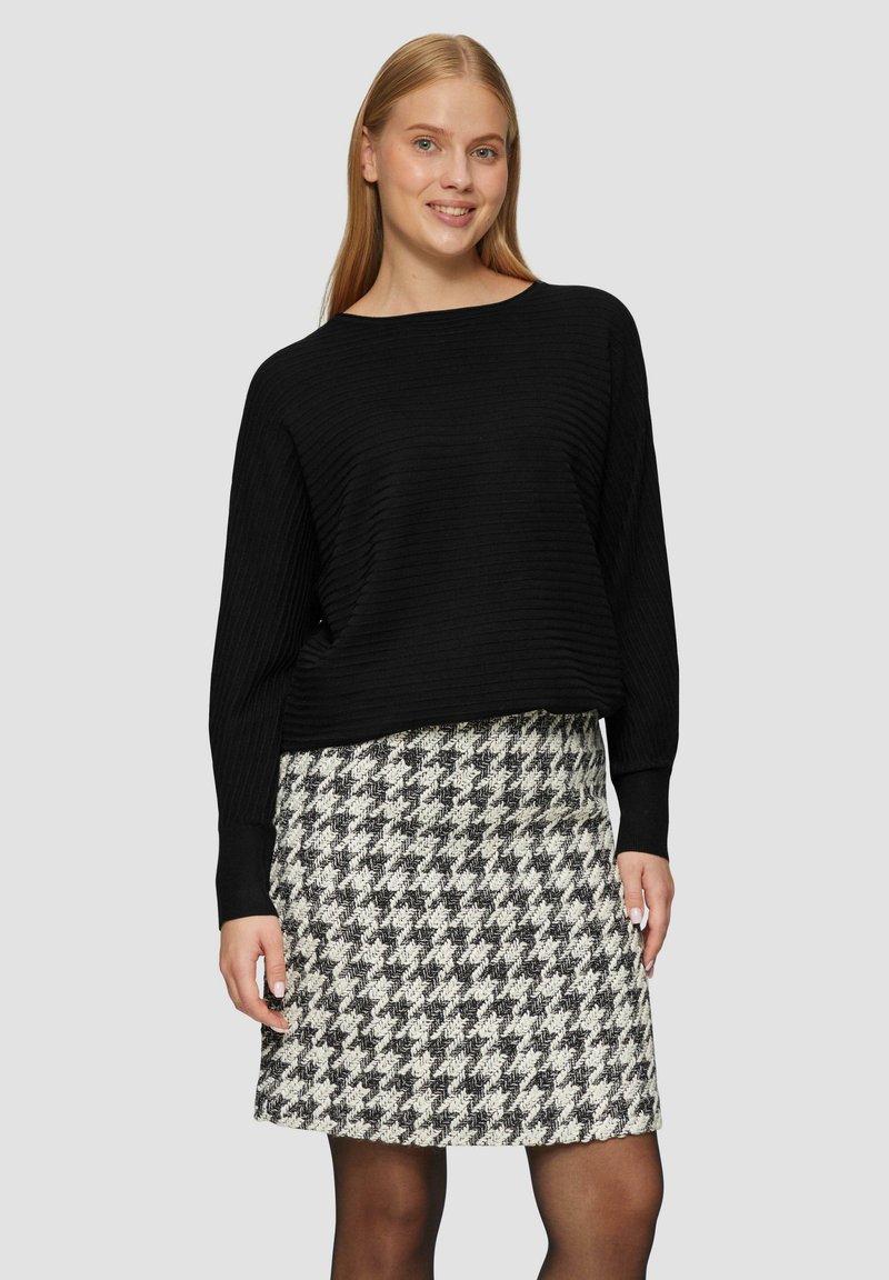 Schwarzer, gerippter Strickpullover mit lockerer Passform, kombiniert mit einem knielangen Rock im Schwarz-Weiß-Hahnentritt-Muster. Das Outfit hat lange Ärmel.