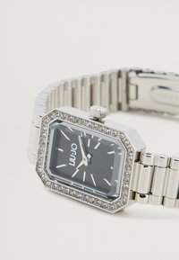 Οκταγωνικός αργυρός ρολόι με μαύρη επιφάνεια, κρυστάλλινες λεπτομέρειες στη στεφάνη και μεταλλικό μπρασελέ. Διαθέτει ασημένιες δείκτες και ωροδείκτες.