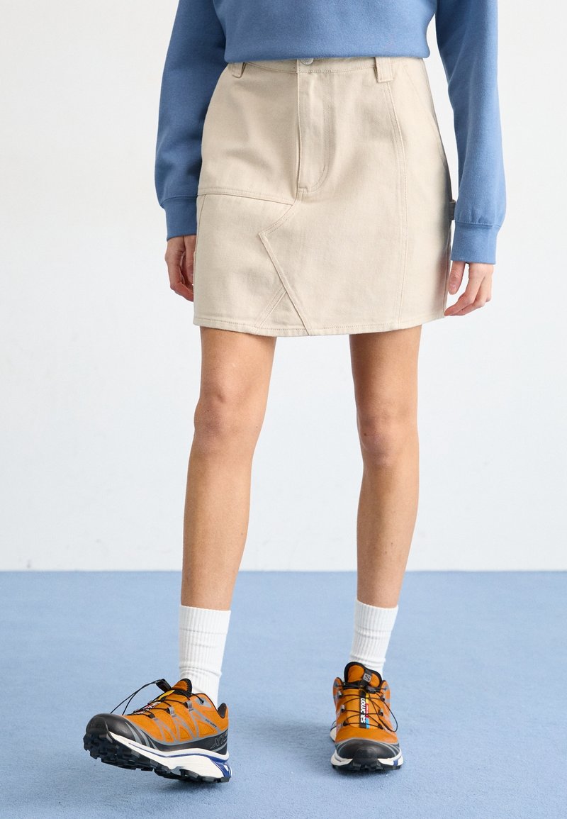 Beige denimkjol med vinklad sömdetalj, ihopparad med en oversized blå sweatshirt och orangea sportskor med svarta detaljer samt vita strumpor.