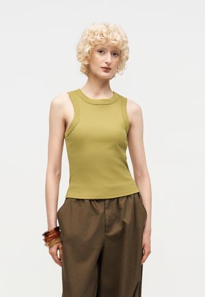 Osoba z kręconymi blond włosami, ubrana w bezrękawnik w kolorze oliwkowej zieleni, brązowe spodnie i bransoletki na lewym nadgarstku, stoi na białym tle.