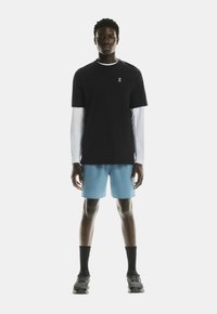Camiseta negra de manga corta sobre una camisa blanca de manga larga, combinada con pantalones cortos de un azul claro. Usada con calcetines negros y zapatillas de correr.
