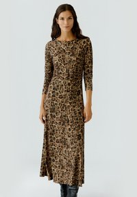 Vrouw in een lange mouw, enkellange jurk met luipaardprint, met zwarte kniehoge laarzen, staand tegen een effen lichte achtergrond.