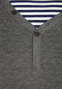 Camicia grigia con colletto a V e bottoni, indossata sopra una maglietta a righe blu e bianche, con particolare attenzione ai dettagli del collo e dei bottoni.