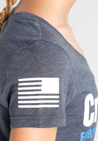 Šedé bavlněné tričko s krátkým rukávem, bílé grafiky vlajky na rameni a modrý text na přední straně. Měkká textura, ležérní design.