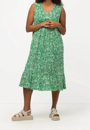 Vrouw draagt een mouwloze groene jurk met wit bloemenpatroon en beige platformsandalen, staand tegen een effen witte achtergrond.