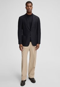 Granatowa marynarka z dwoma przednimi kieszeniami, noszona na czarnym golfie; zestawiona z beżowymi prążkowymi spodniami i czarnymi butami. Gładka faktura tkaniny.