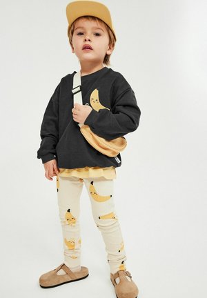 Jong kind met een gele pet, zwart sweatshirt met lachende banaan, crèmekleurige broek met banaan prints, beige tas en bruine schoenen, staand tegen een witte achtergrond.