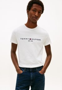 Noor mees, kellel on käed taskutes, kannab valget Tommy Hilfigeri t-särki ja tumedasiniseid teksaseid ühtlase heleda tausta ees.