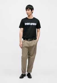 Άνδρας που στέκεται φορώντας μαύρο T-shirt "DONDUP", μπεζ πλισέ παντελόνια με μαύρη ζώνη, μαύρα παπούτσια, με τα χέρια στις τσέπες, μπροστά σε απλό φόντο.