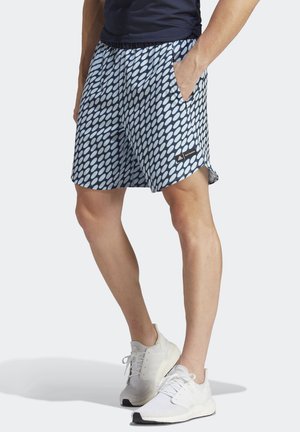 Person iført marineblå og hvide mønstrede shorts med hånden i lommen og hvide sneakers, stående mod en lys baggrund.