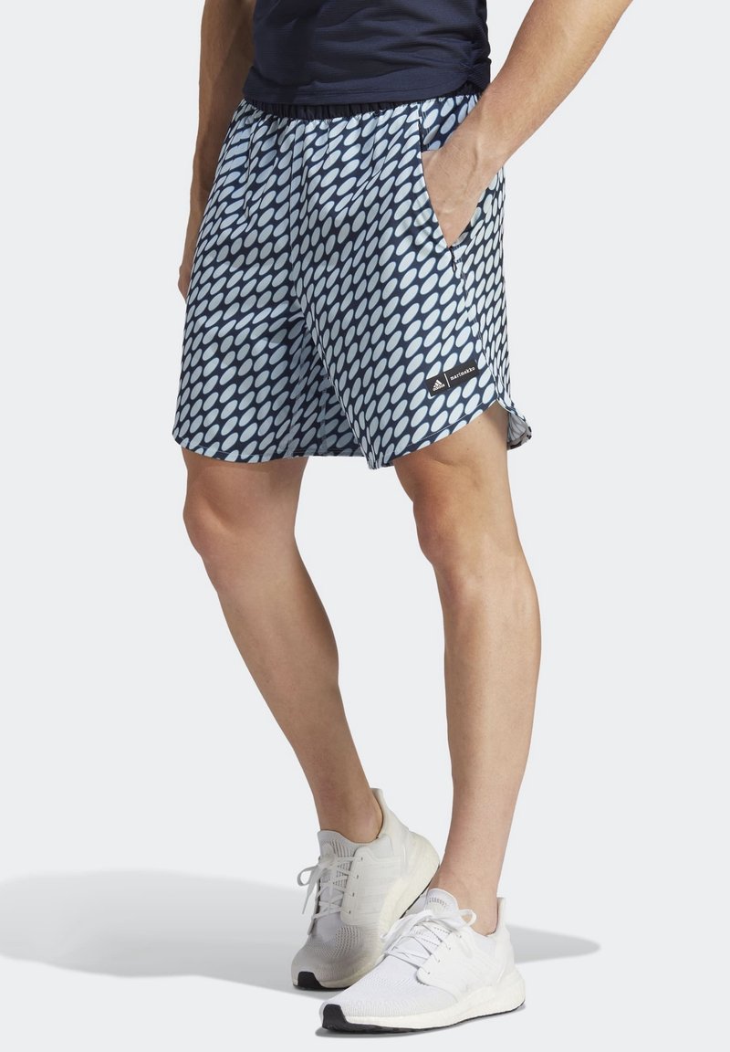 Personne portant un short à motifs bleu marine et blanc, la main dans la poche et des baskets blanches, debout devant un fond clair.
