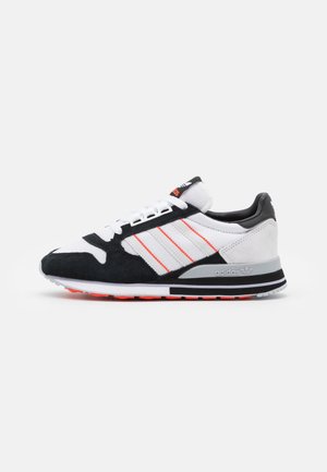 Scarpa da ginnastica Adidas bianca e nera con tre strisce laterali rosse, lacci bianchi e suola grigia con dettagli neri e rossi.