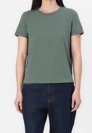 Osoba nosząca zielony t-shirt z krótkim rękawem i okrągłym dekoltem, zestawiony z ciemnoniebieskimi dżinsami, widoczna od barków do bioder.