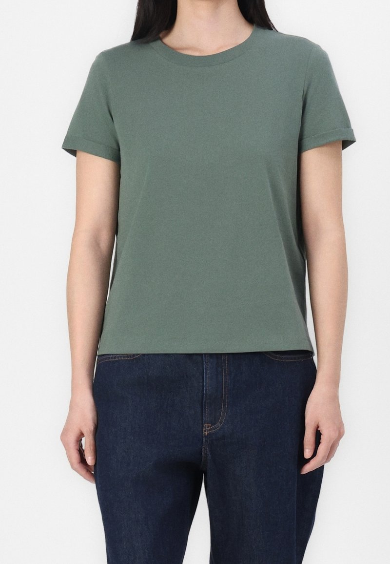 Personne portant un t-shirt vert à manches courtes avec col rond, associé à un jean bleu foncé, visible des épaules jusqu'aux hanches.