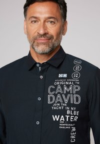 Camisa negra con diseño abotonado, que presenta gráficos de texto en blanco y de color. Tejido de algodón con un acabado texturizado y botones en contraste.