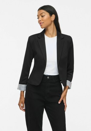 Femme portant un blazer noir avec des poignets rayés retroussés, une chemise blanche et un pantalon noir, regardant vers sa gauche sur un fond clair uni.