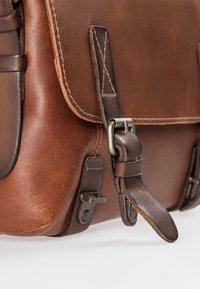 Borsa a tracolla in pelle marrone con finitura texturizzata, cinturino regolabile, accessori in metallo e cuciture dettagliate sul cappuccio e sui lati.