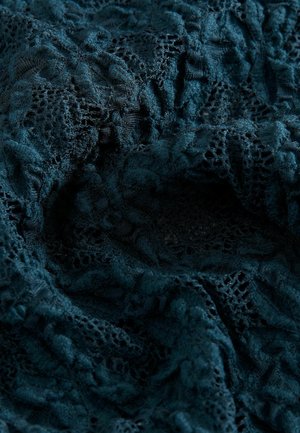 Mörk teal spetstyg med intrikata mönster och en mjuk, texturerad yta som visar varierande grad av genomskinlighet och detaljerad broderi.