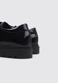 Zapatos de charol negros con punta redonda, acabado suave y una suela de goma negra gruesa con diseño de relieve.