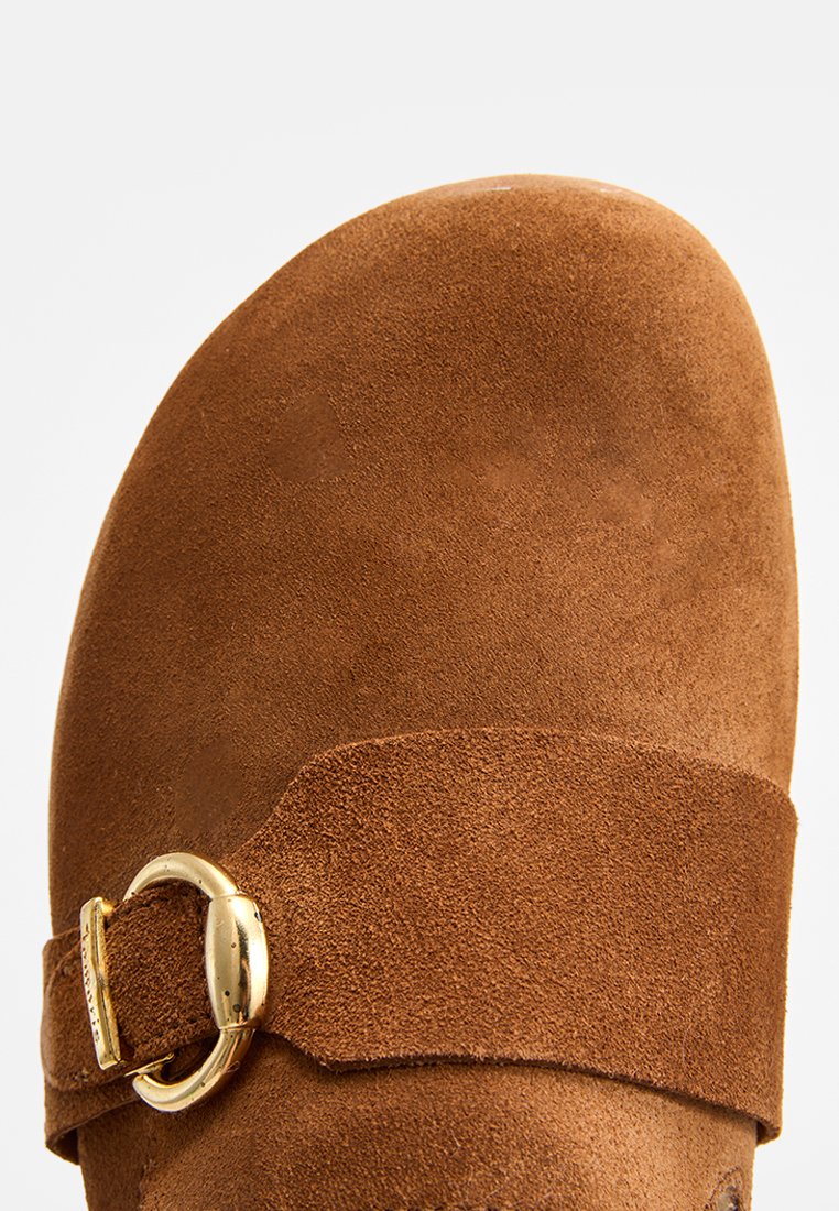 Bruine suede pantoffel met brede band en gouden gespdetail. Afgeronde neus en zachte textuur, ideaal voor comfort en casual draag.