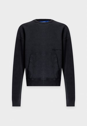 Jeune homme portant un pull oversize foncé, un pantalon noir et des baskets noires à semelles blanches, debout devant un fond clair uni.