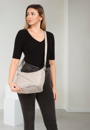 Vrouw met zwarte top en donkere spijkerbroek draagt lichtgrijze crossbodytas met verstelbare band in een neutrale binnensetting.